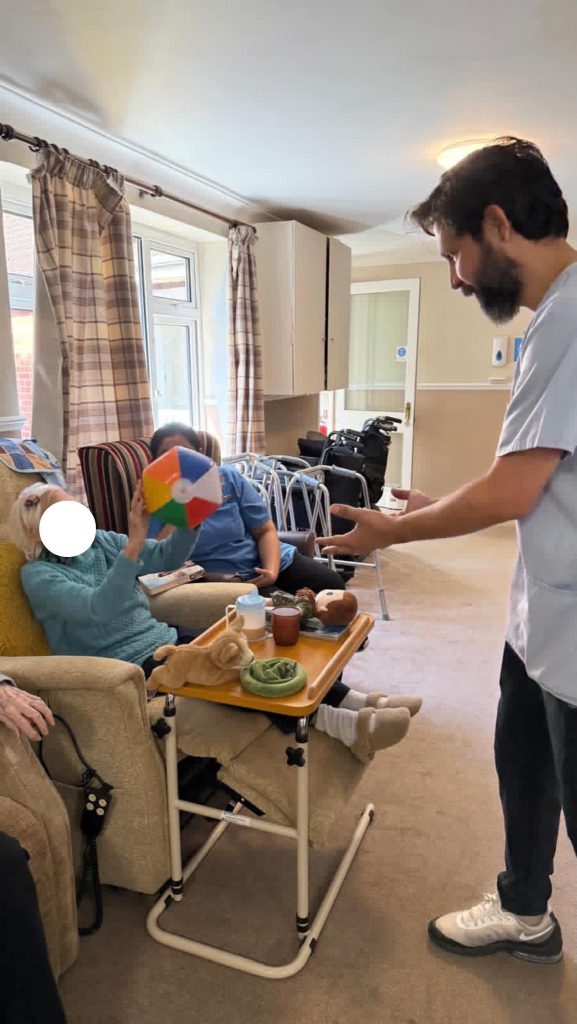 An elderly woman sits in a chair holding a colorful ball, reaching toward a caregiver standing nearby.