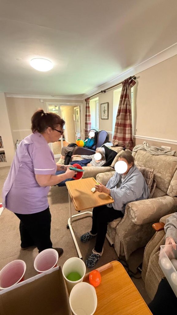 Caregiver assisting elderly residents in a lounge as they play a simple cup-and-ball game together.