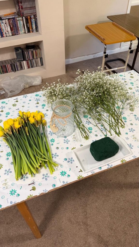 Table with yellow flowers, greenery, a glass vase, and floral foam.