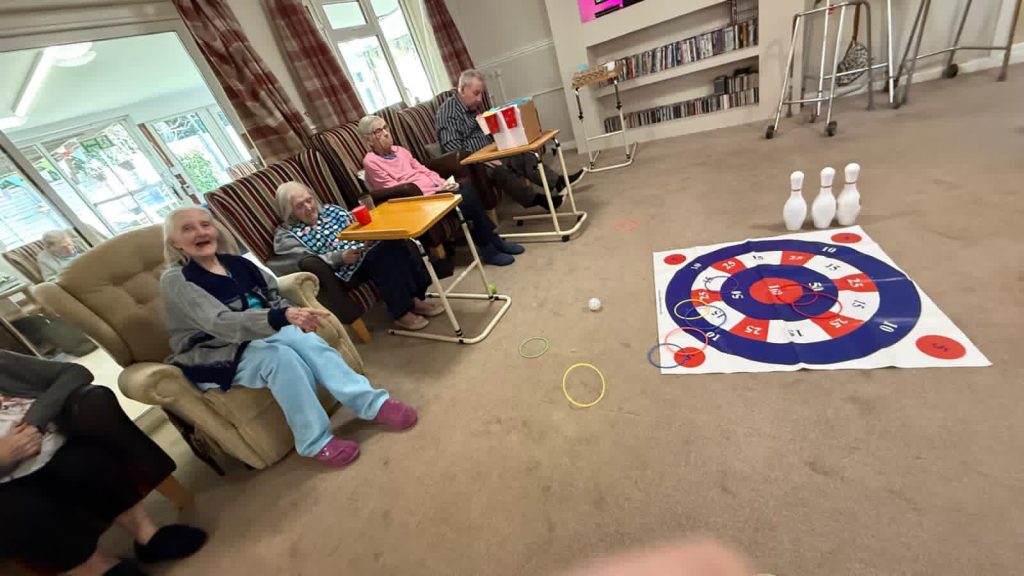 A group of elderly people sit in a lounge, playing a bowling-style game with rings and a target mat on the floor.
