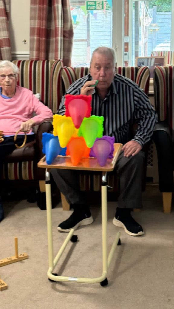 An older man sits at a small table, blowing into a device toward a stack of colorful balloon-like shapes, while an elderly woman sits beside him watching.