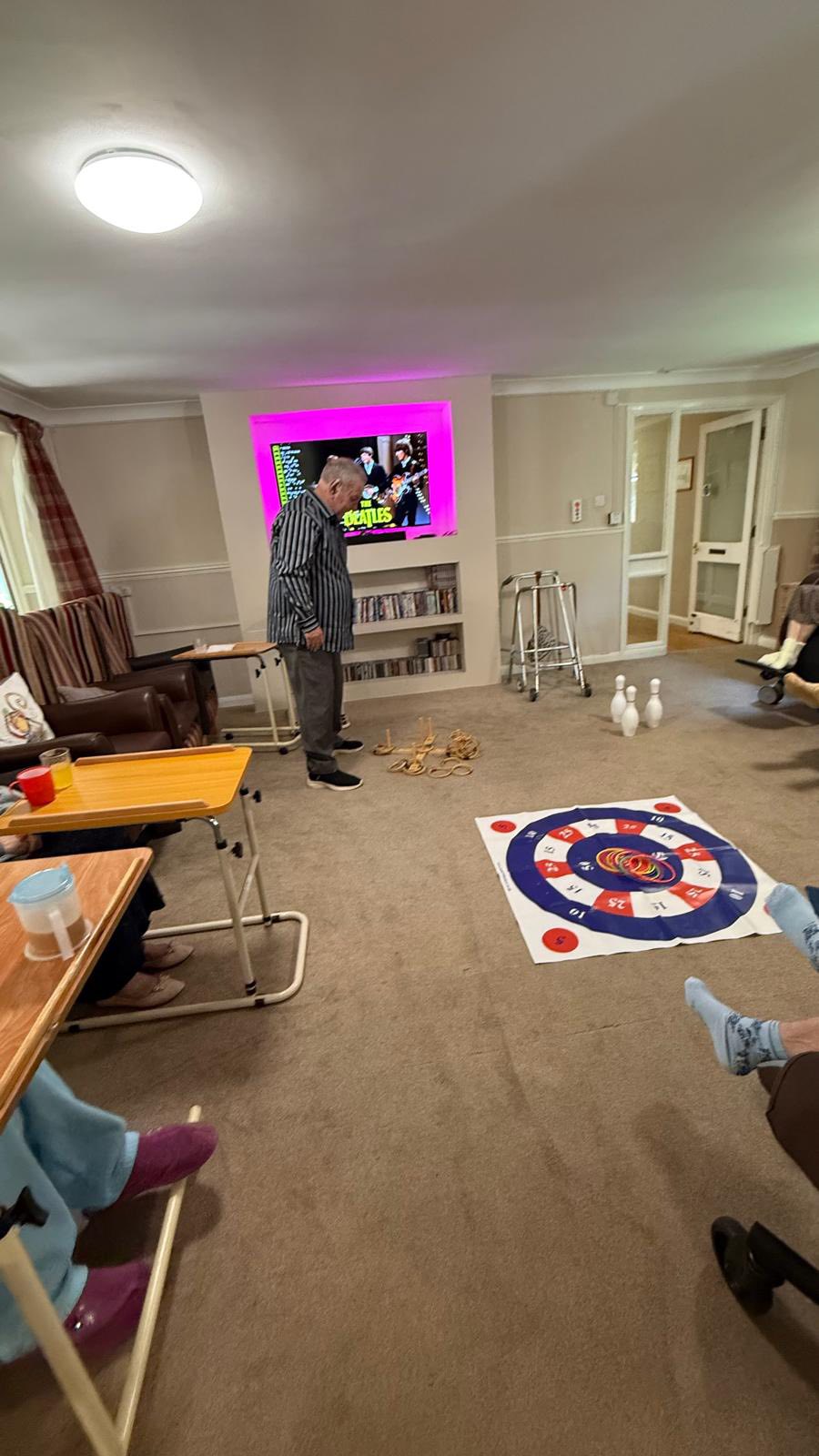 Elderly people in a lounge playing indoor games, including ring toss and floor target, with a man standing and preparing to throw.