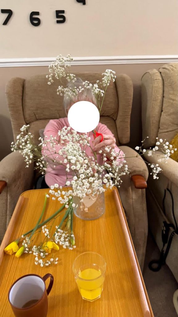 Elderly woman sitting with a finished flower arrangement in a glass jar.