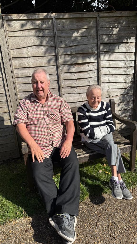 Older man and woman sitting on a bench outdoors with a wooden fence behind them