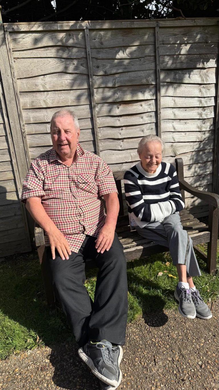 Older man and woman sitting on a bench outdoors with a wooden fence behind them