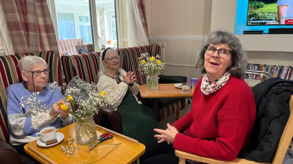 Elderly woman arranging flowers on a tray with yellow and white blooms.