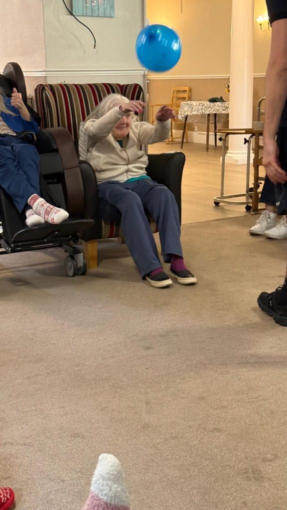 Residents seated in a lounge, with some holding or reaching for balloons during a group activity