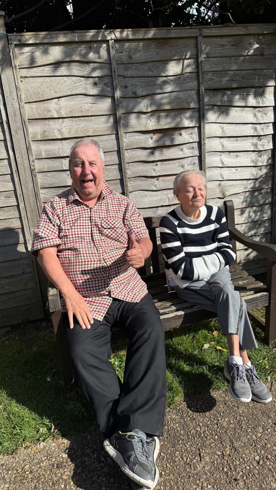 Man and woman sitting outdoors on a bench, with a wooden fence in the background
