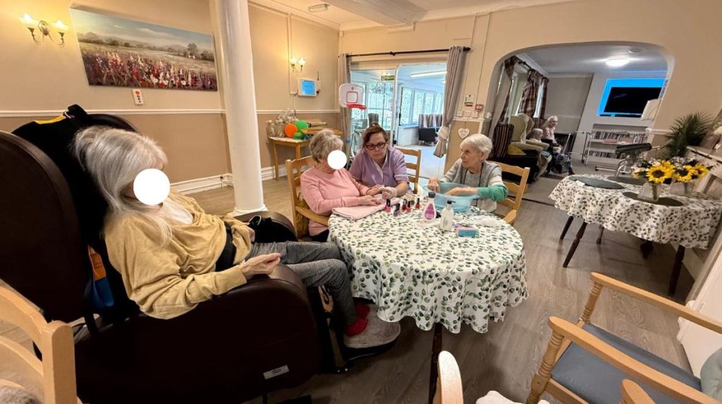 Group of elderly women sitting together while a care worker helps with nail painting.