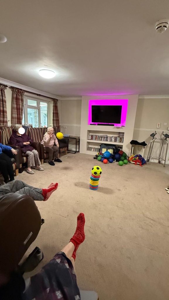 Elderly residents sitting in a circle playing a ball game in a lounge.