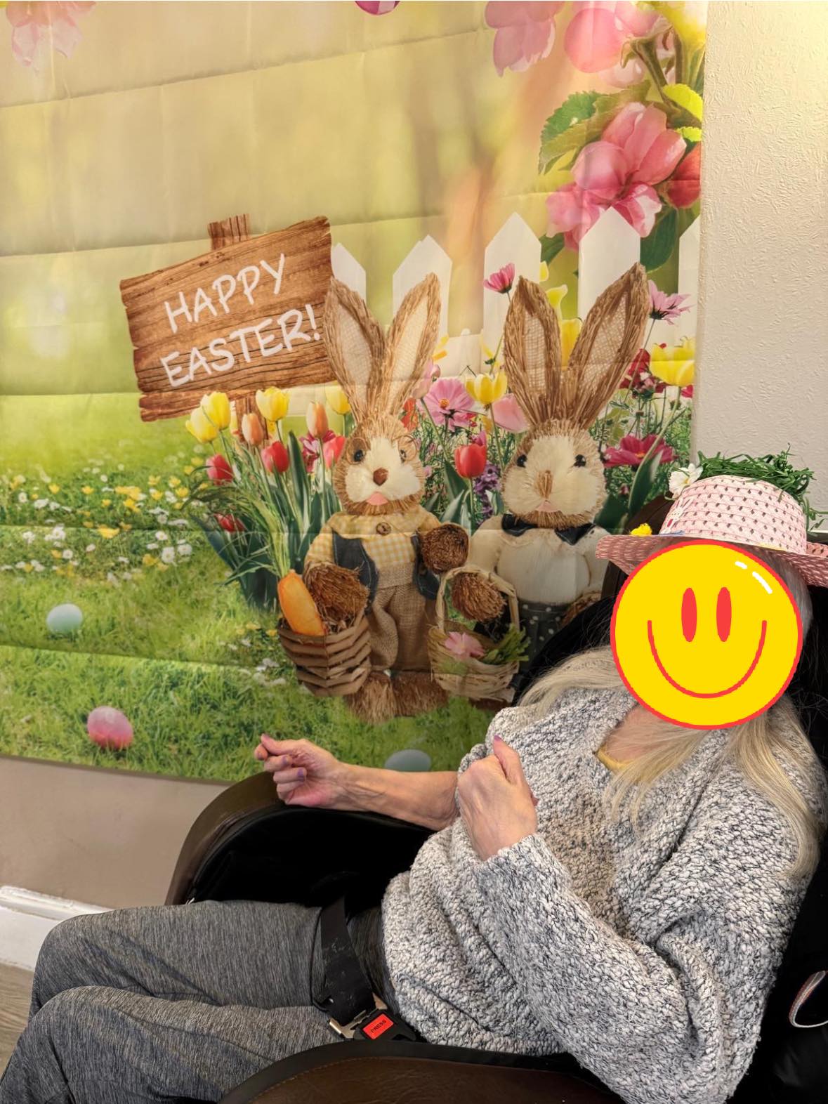 Person sitting beside a “Happy Easter” backdrop with bunny figures, wearing a pink decorated hat.