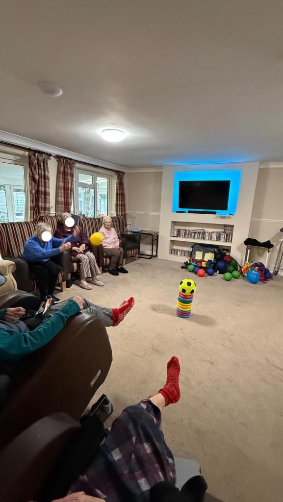 Group of elderly residents playing a seated ball game in a lounge area.