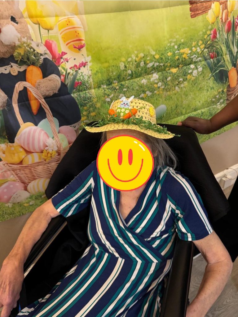 Elderly woman in a striped dress wearing a decorated Easter hat, seated in a chair with a festive spring backdrop.