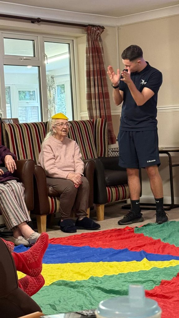 Care worker clapping while an elderly woman sits wearing a small cone hat during a group activity.