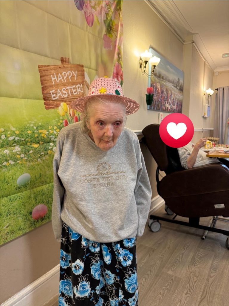Elderly woman wearing a pink hat stands in front of an Easter-themed backdrop in a care home.