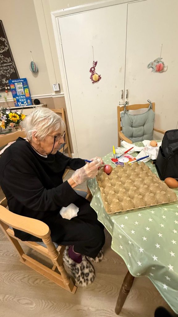 Elderly woman painting eggs at a table in a care home.