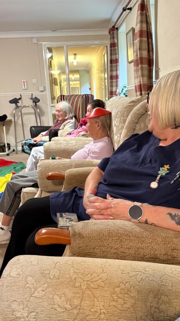 Elderly residents seated in a row, some wearing cone hats, watching the activity.
