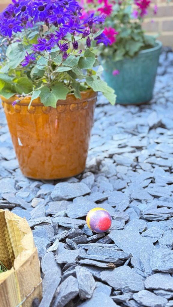 Bright painted egg on gravel near vibrant potted flowers.