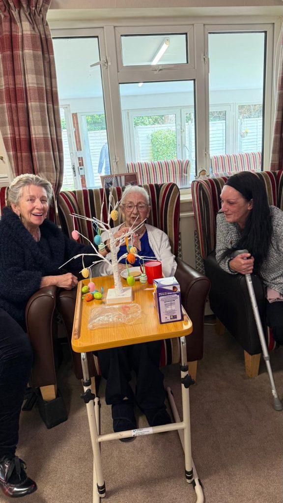 Elderly woman with two companions decorating Easter egg tree in a care home.