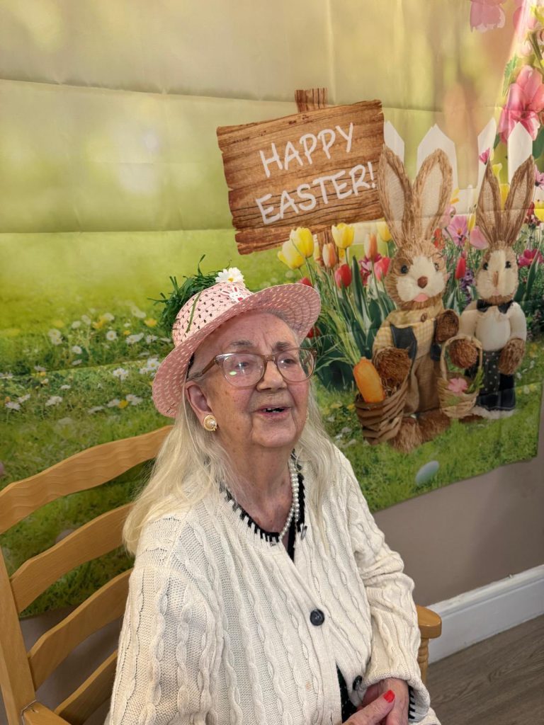 Elderly woman smiling in Easter hat in front of festive backdrop.