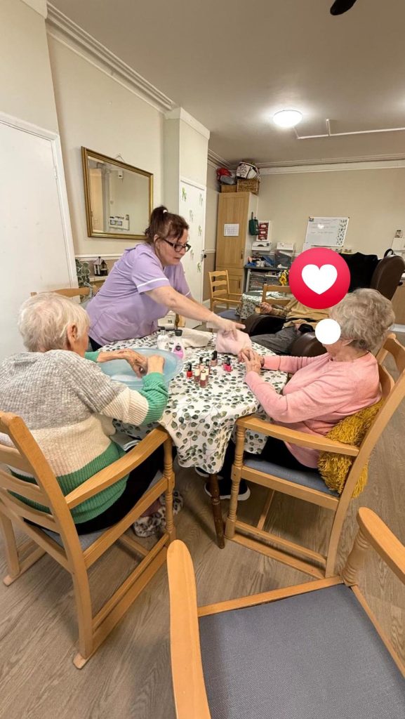 Care worker helping two elderly women paint their nails at a table.