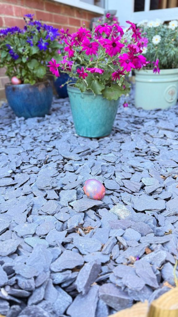 Pink and purple flowers in pots with a painted egg on gravel.