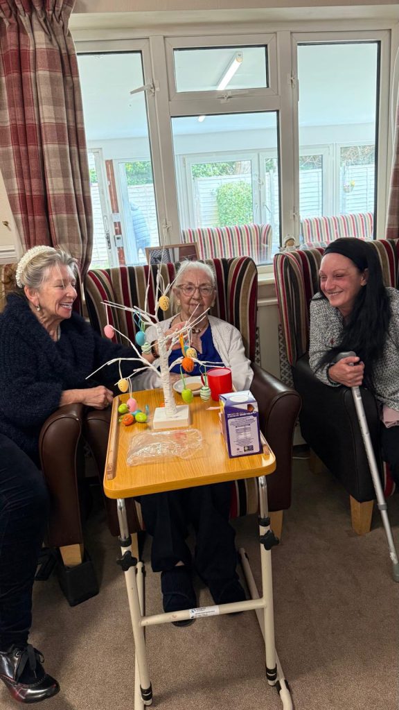 Three women smiling and decorating Easter egg tree together indoors.