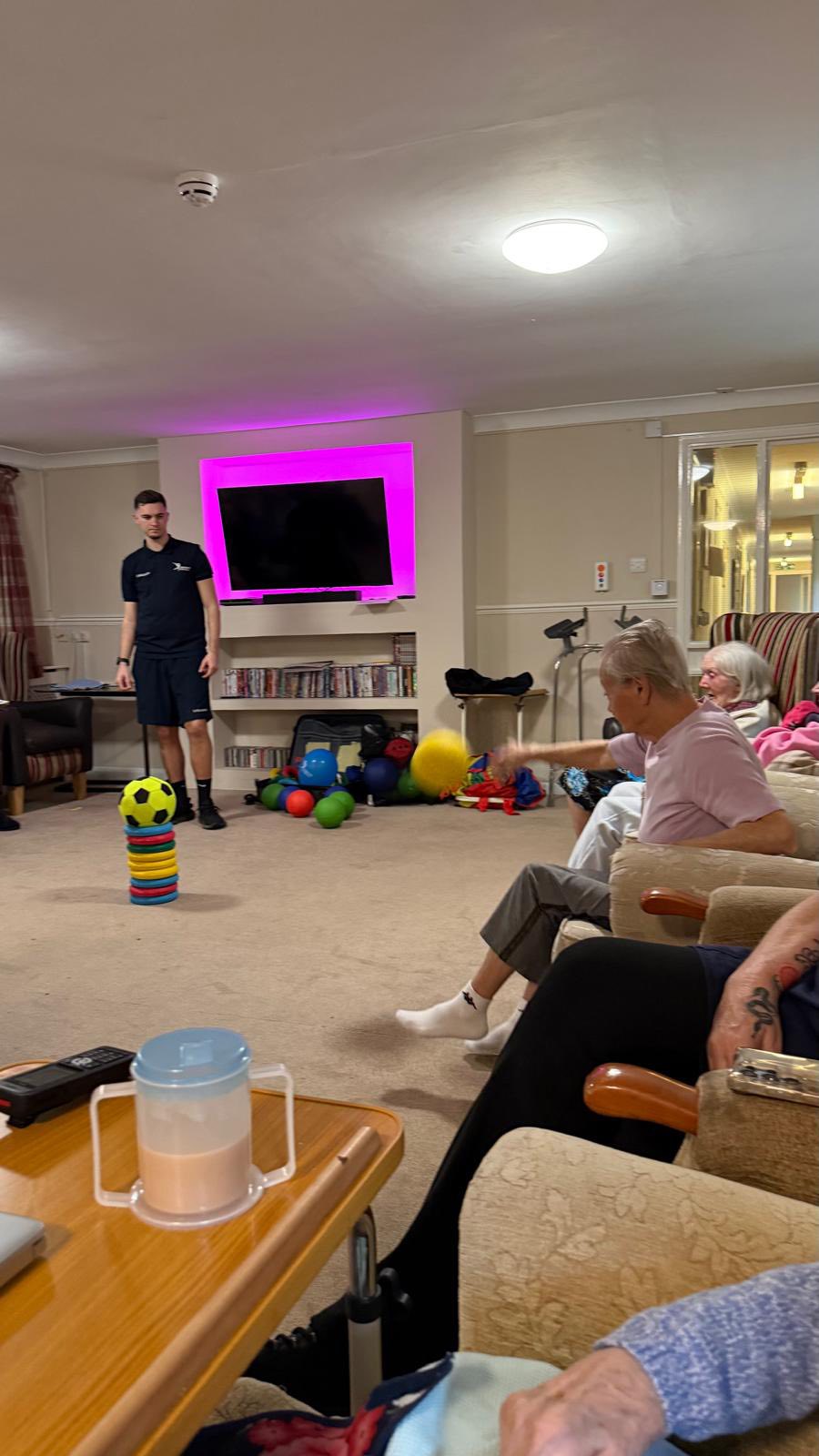 Elderly woman throwing a yellow ball toward a target while others watch.