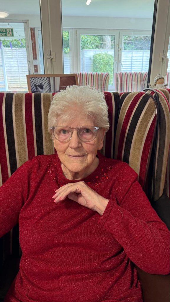 Elderly woman in a red sweater sitting in a chair, smiling at the camera.