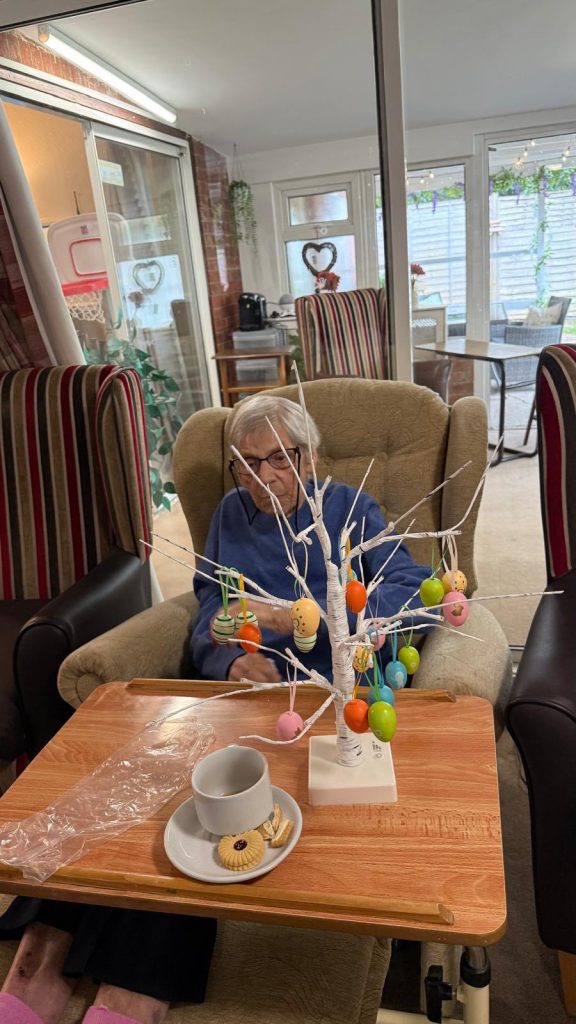 Senior woman sitting in armchair decorating a small Easter egg display tree.