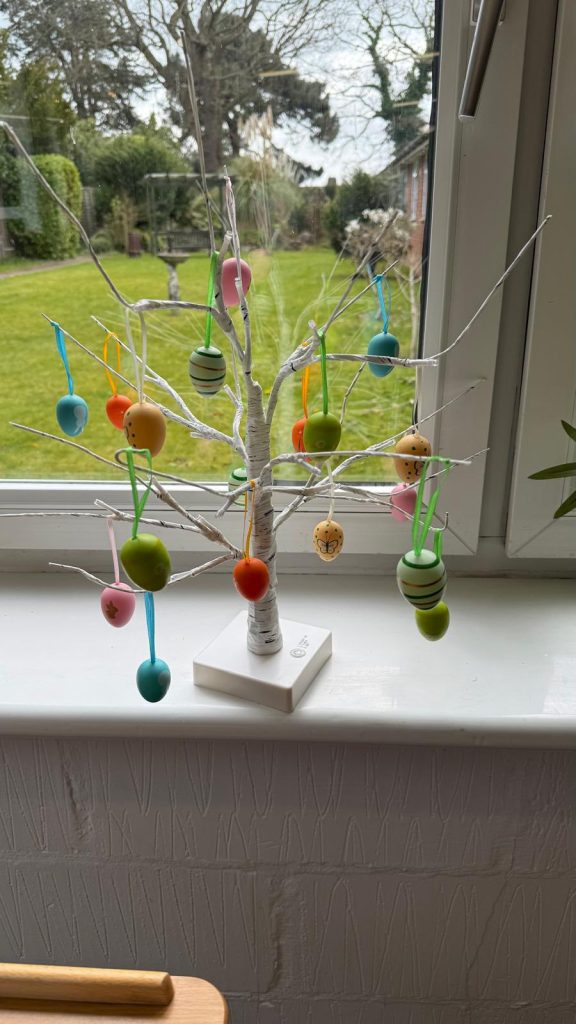 Colorful Easter eggs hanging on a decorative white tree by a window.