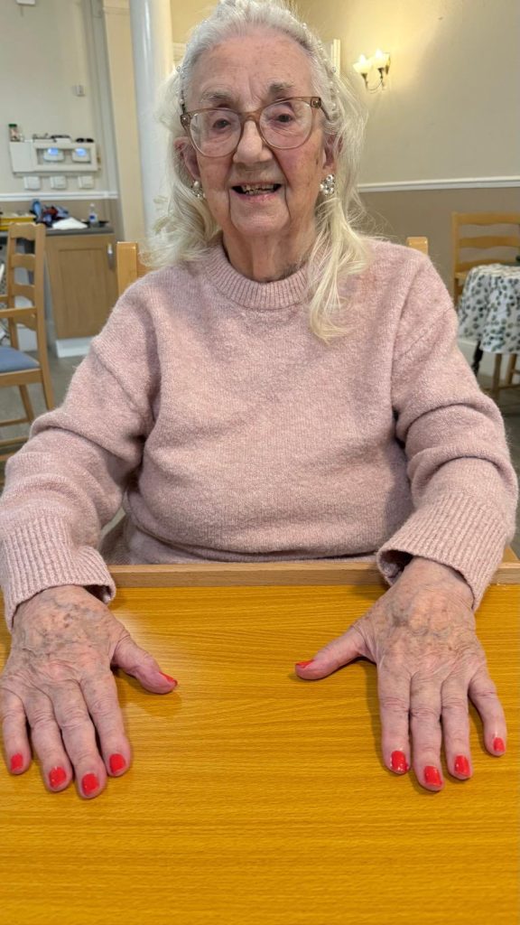 Elderly woman in a pink sweater sitting at a table with red painted nails.