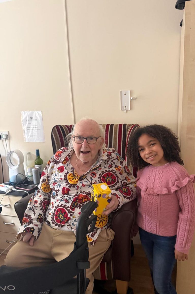 An elderly man sits smiling in a chair holding a small box, with a young girl standing beside him smiling.
