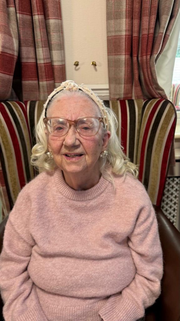 Elderly woman in a pink sweater sitting in a chair, smiling gently.