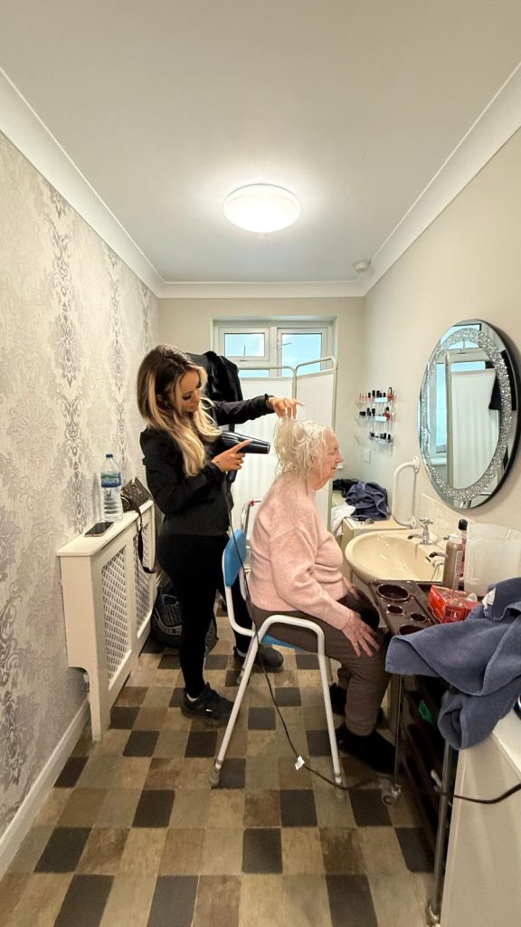Care worker drying an elderly woman’s hair in a small salon area.