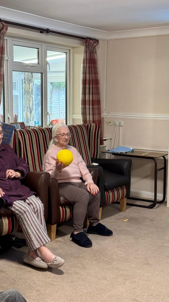 Elderly woman holding a yellow ball while seated in a chair.