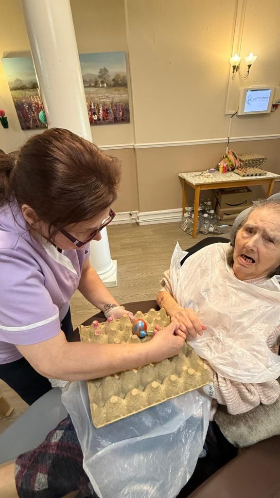 Caregiver helping elderly woman paint an Easter egg during activity session. 