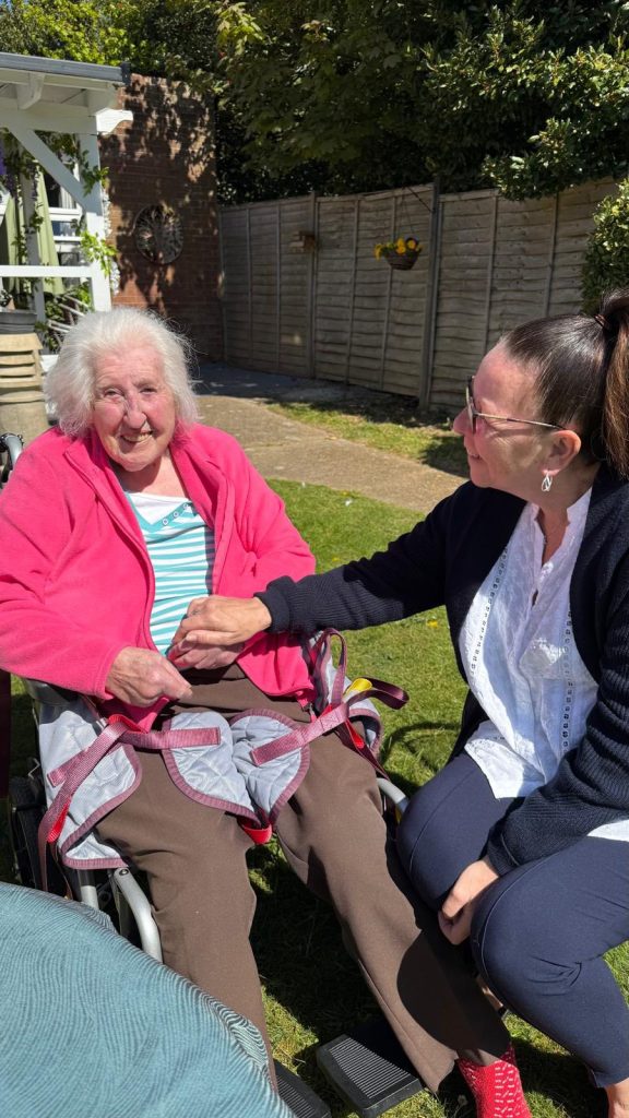 Elderly woman in a wheelchair smiling while holding hands with a caregiver in a sunny garden.