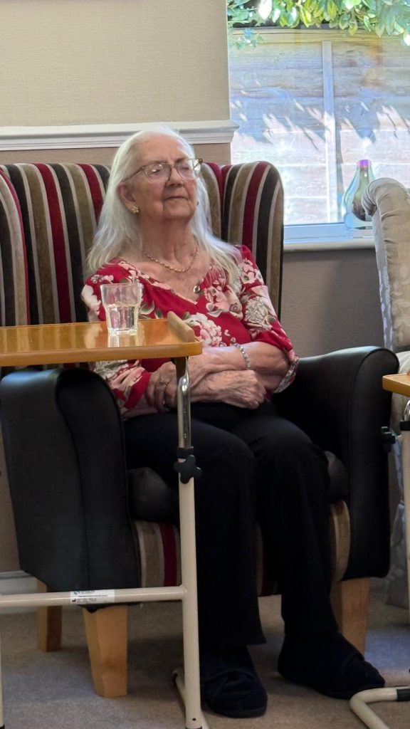 Elderly woman with white hair sitting calmly in an armchair by a window.