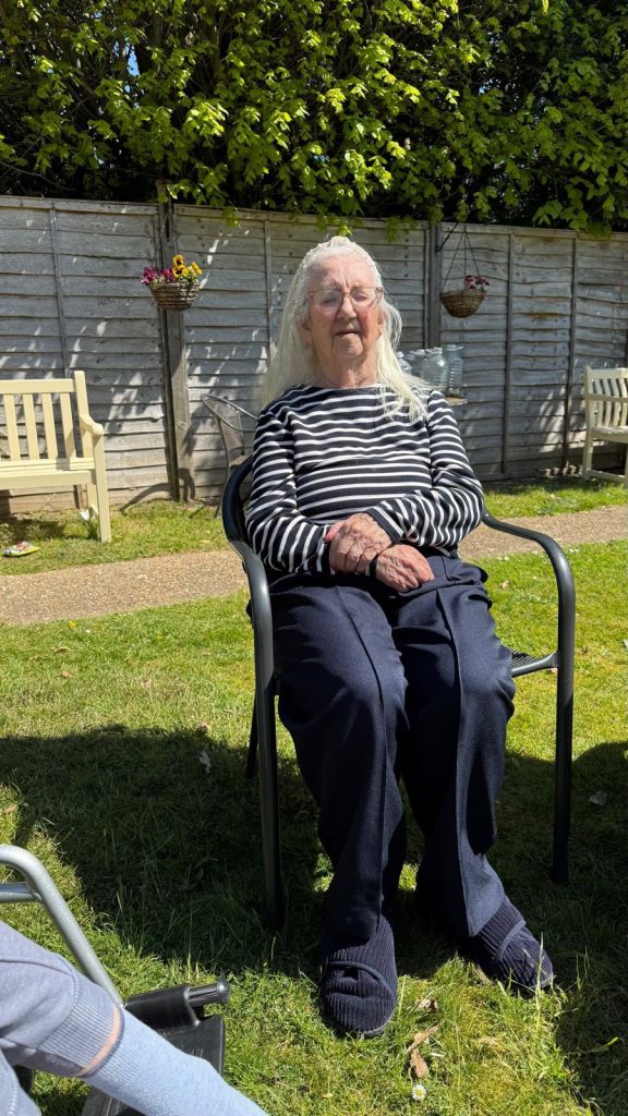 Elderly woman sitting calmly on a chair in a sunny garden setting.