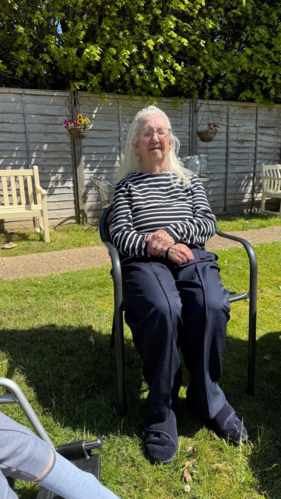 Elderly woman sitting calmly on a chair in a sunny garden setting.