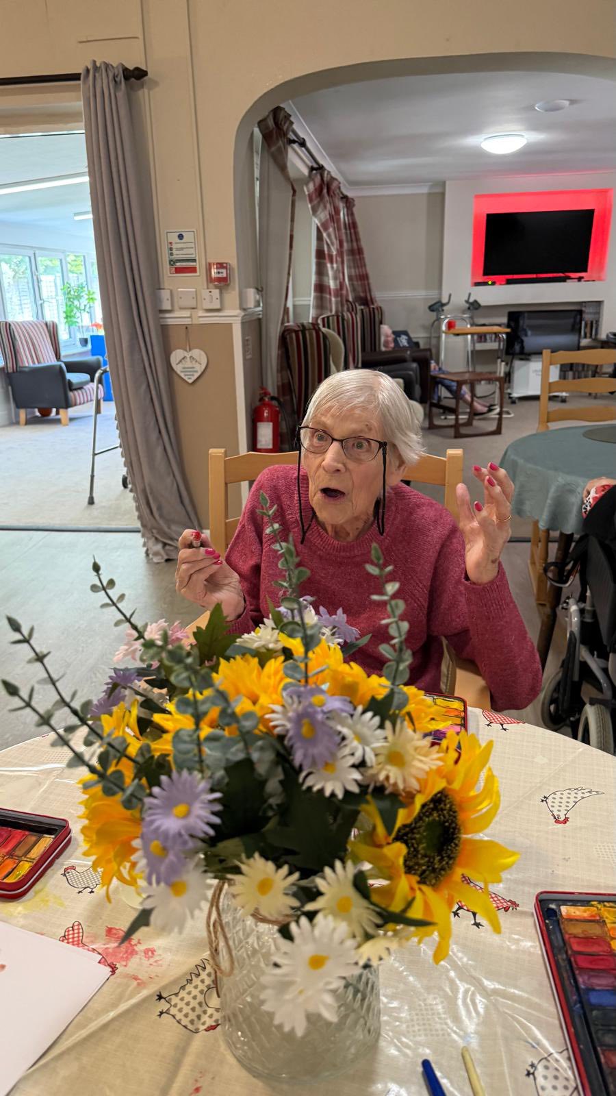 Elderly woman reacting happily while painting, with a vase of flowers in the foreground