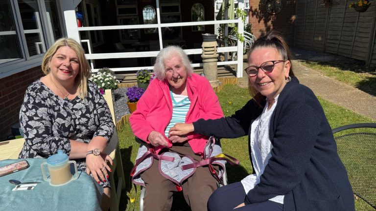 Elderly woman in a wheelchair smiling with two caregivers seated beside her in a bright outdoor garden setting.