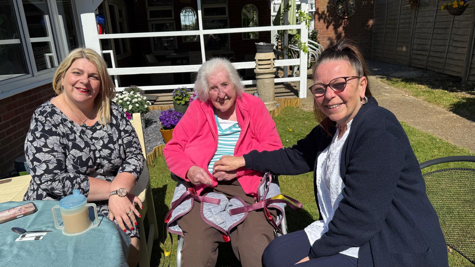 Elderly woman in a wheelchair smiling with two caregivers seated beside her in a bright outdoor garden setting.