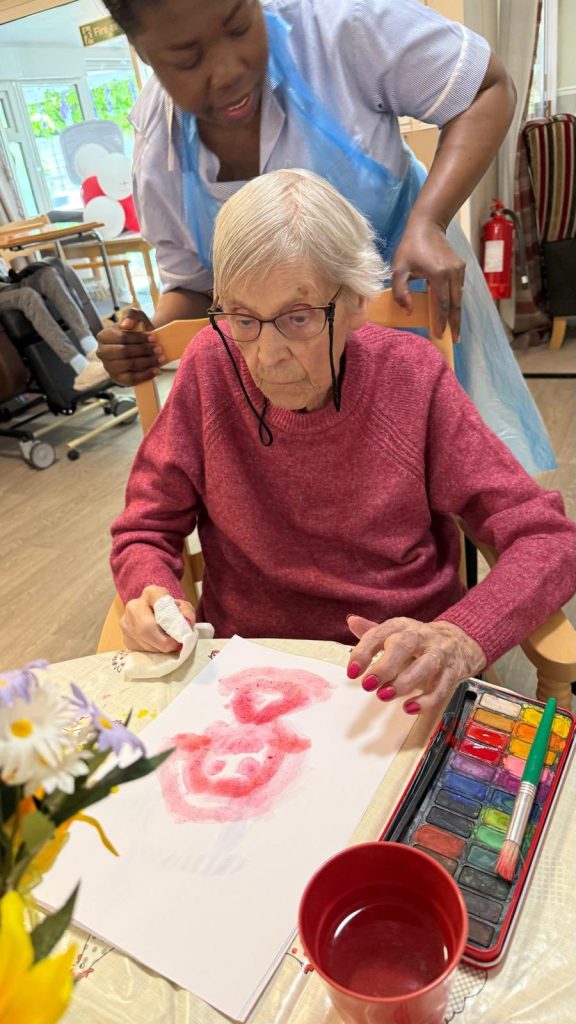 Elderly woman painting with watercolors while a caregiver supports from behind.