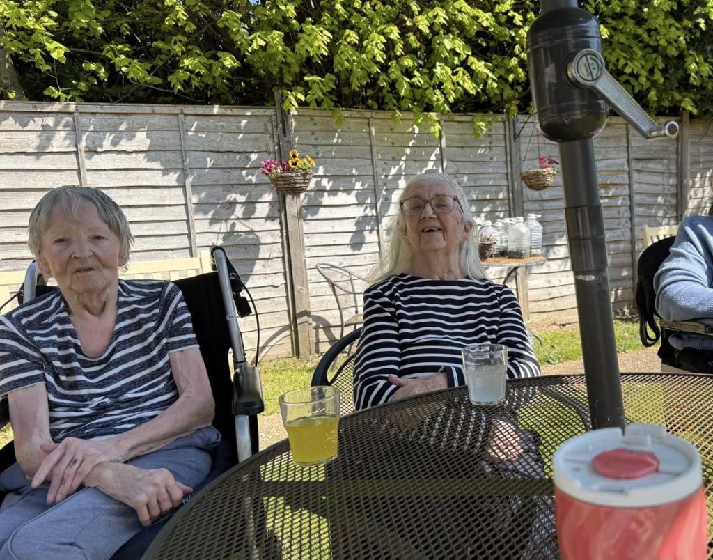Elderly women sitting outdoors at a garden table enjoying drinks on a sunny day.