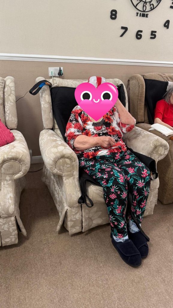 Elderly woman seated in a recliner chair wearing patterned clothes and slippers.