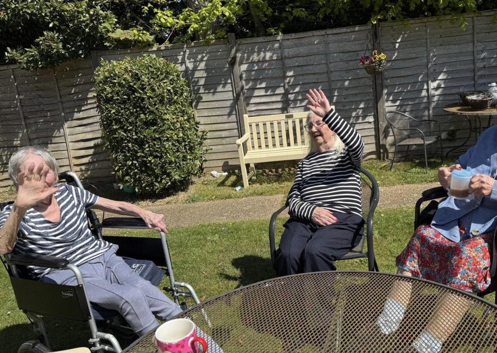 Two elderly women in a garden raising hands playfully while sitting outside.