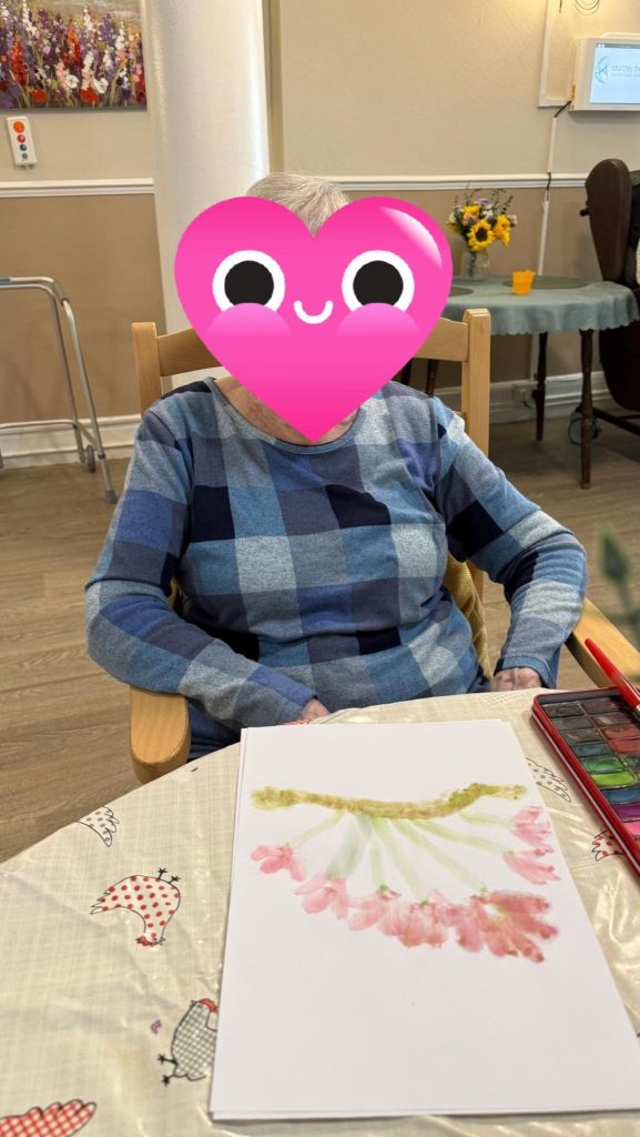 Elderly woman seated with a finished flower painting on paper in front of her.