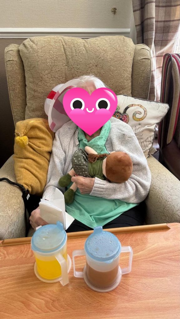 Elderly woman sitting in a chair holding a doll, with drinks on a table.
